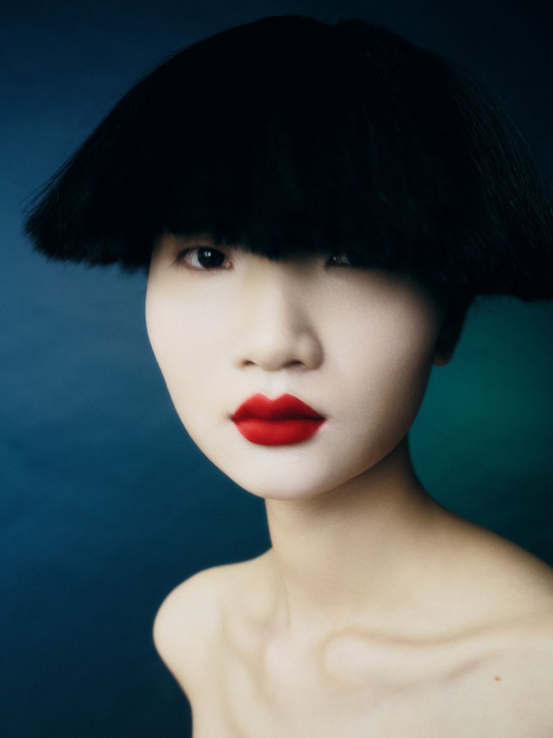 Close-up portrait of a model with short black bob hair, pale complexion, and vibrant red lipstick against a dark blue background.