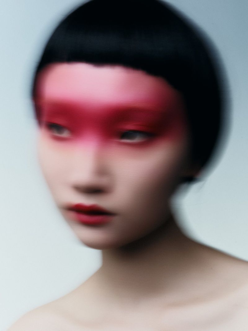 Close-up beauty portrait of an East Asian model with short black hair and blurred red makeup across the eyes against a pale backdrop.
