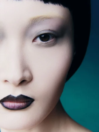 Close-up beauty portrait of an East Asian model with bleached eyebrows, blunt black bangs, and dark lined lips against a teal backdrop.