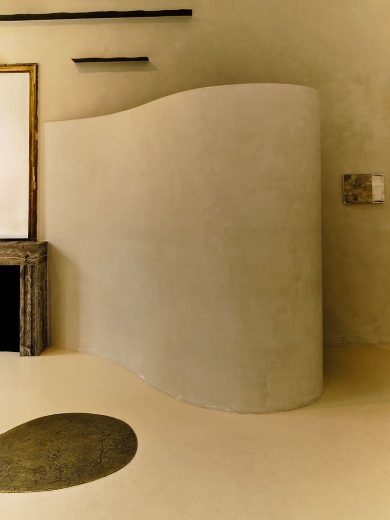 Minimalist interior with a curved beige plaster wall divider, smooth light flooring, and an organic dark circular floor inlay.