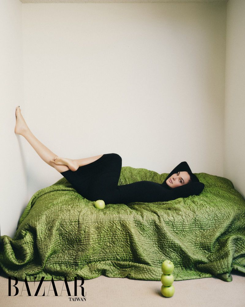 Gal Gadot reclining on a textured green quilt wearing a black hooded dress with one foot propped against a neutral studio wall.