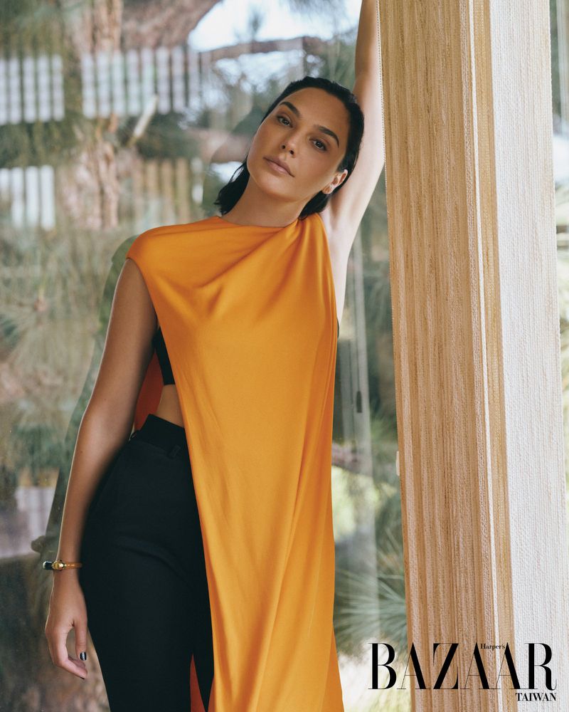 Gal Gadot posing in a draped marigold sleeveless top and black trousers beside a wooden pillar for Harper's Bazaar Taiwan.