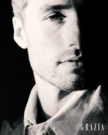 Black and white close-up portrait of a man with light facial stubble wearing a collared shirt and tailored jacket in dramatic lighting.