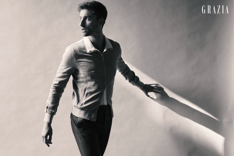 Monochrome fashion portrait of a man in a zip-up collared jacket and dark trousers standing against a pale studio wall