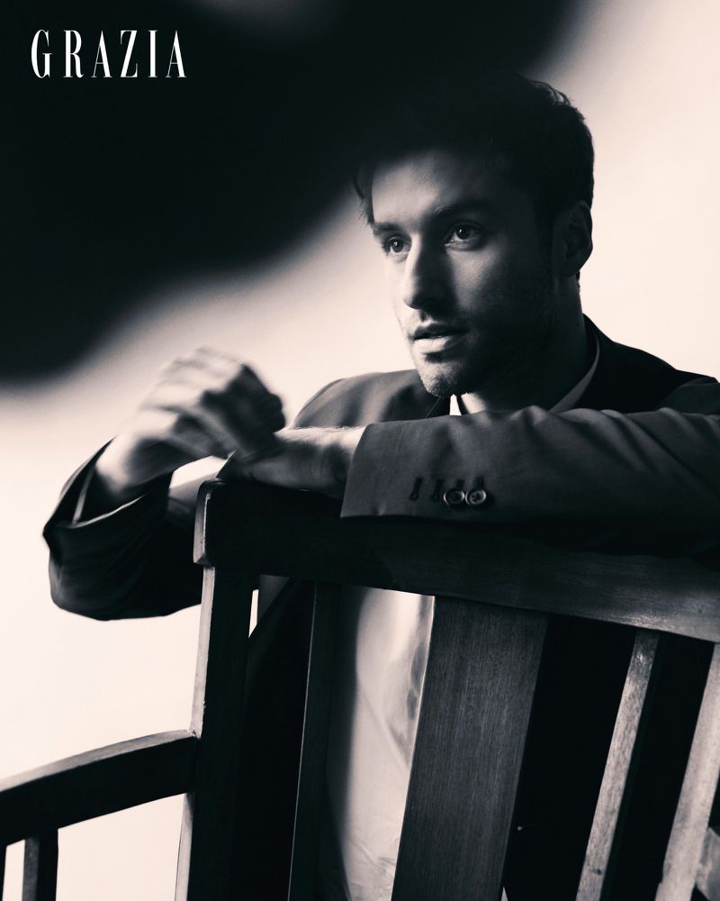 Monochrome portrait of a man in a dark tailored suit jacket leaning over the back of a wooden chair.