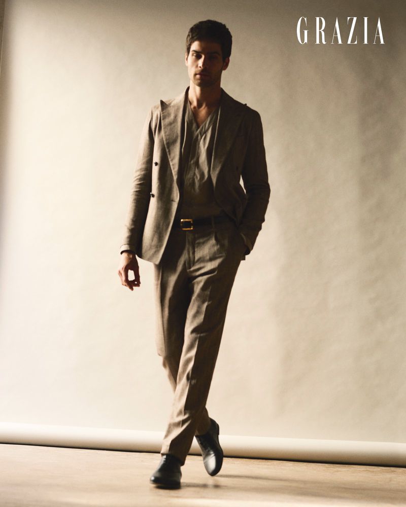Man walking forward wearing a tailored taupe suit and black dress shoes against a warm studio backdrop.