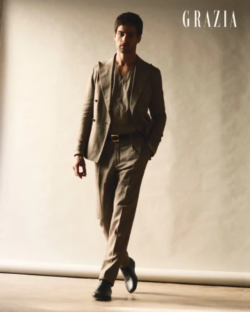 Man walking forward wearing a tailored taupe suit and black dress shoes against a warm studio backdrop.