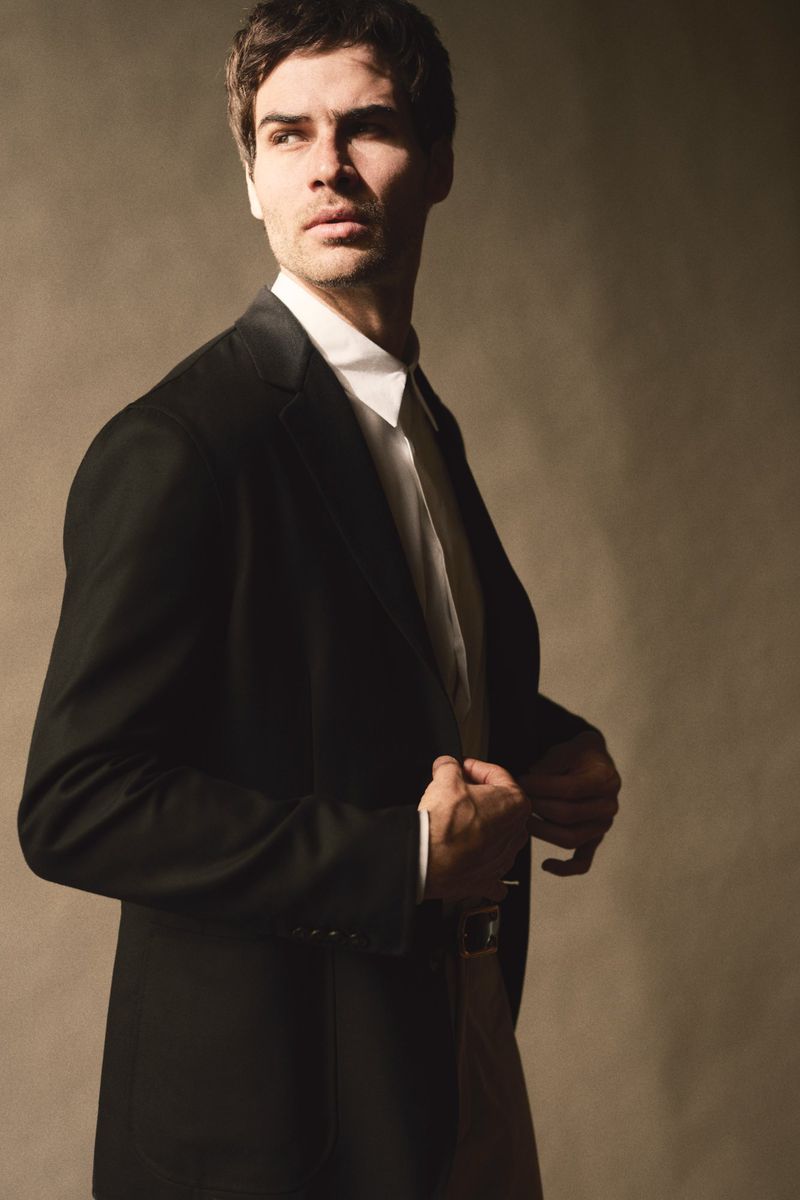 Man in a tailored black blazer and crisp white shirt posing against a warm neutral studio backdrop.