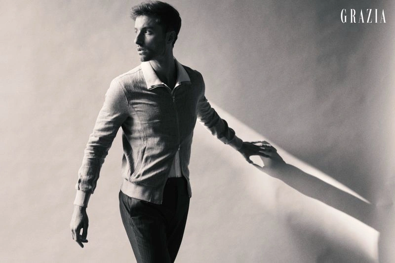 Monochrome fashion portrait of a man in a zip-up collared jacket and dark trousers standing against a pale studio wall