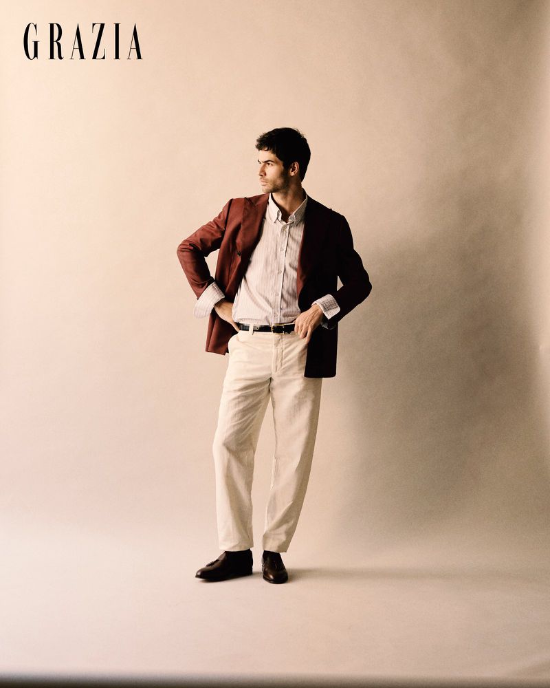 A man posing in a rust-toned blazer, white shirt, and cream trousers against a warm seamless studio backdrop.