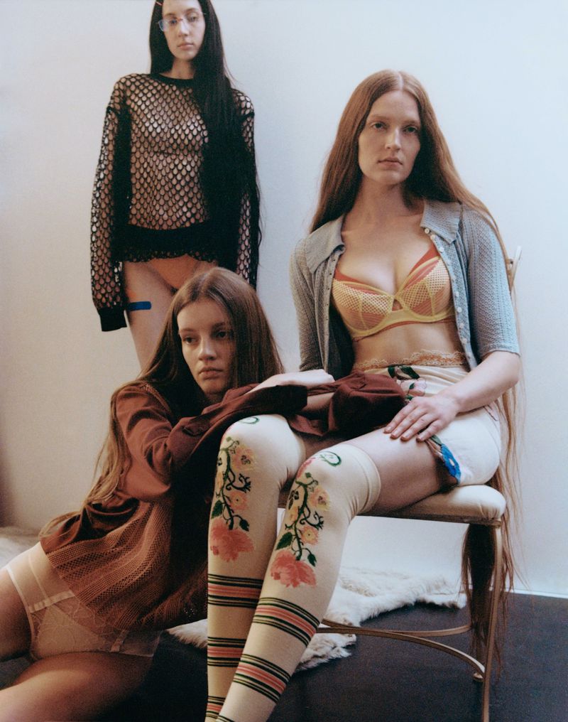 Three women in a photo studio setting. One is seated wearing floral stockings and a yellow bra, with one partially visible and a third standing.