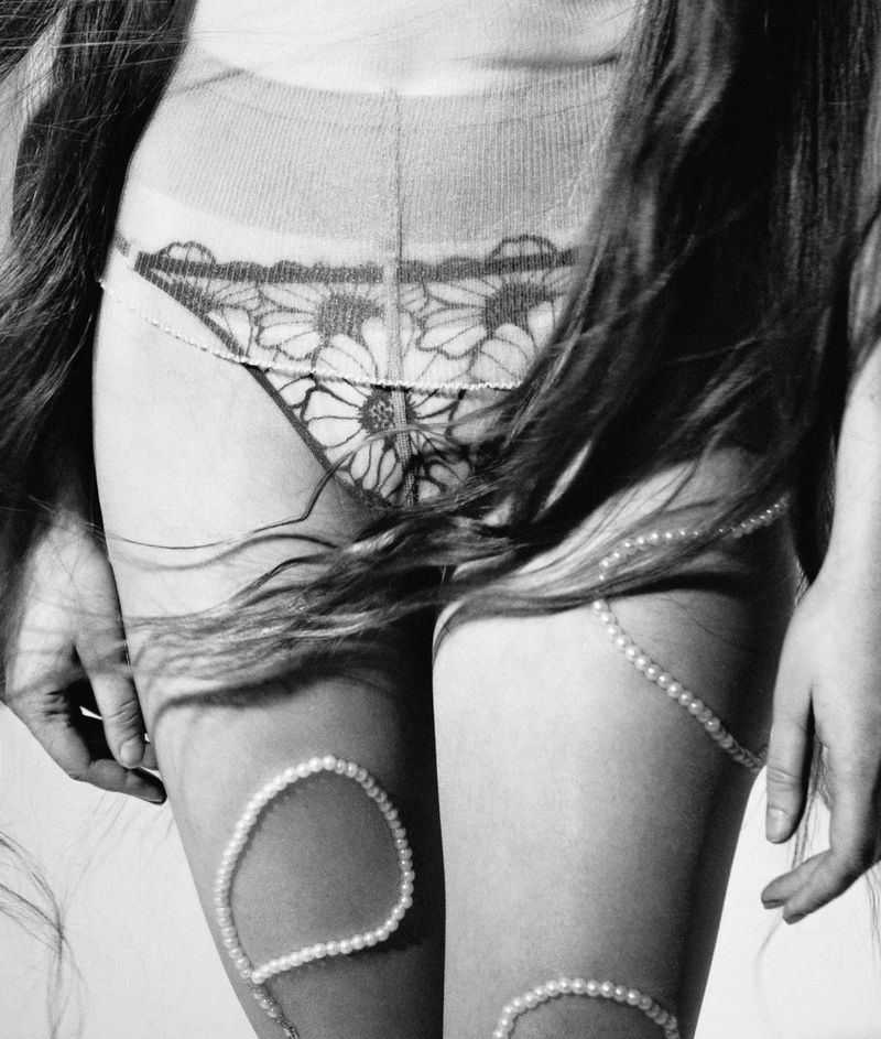 Black and white studio shot featuring a young woman's torso and legs, adorned with a floral panty design and pearl strands.
