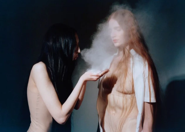 Two women, one with dark hair blowing powder onto another with auburn hair, in a nude-toned setting, creating a hazy atmosphere.