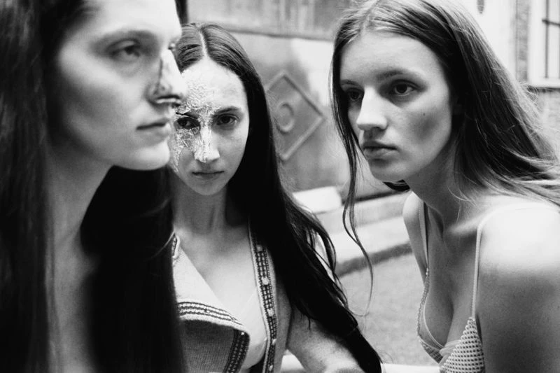 Black and white portrait of three women with long hair, one with partial face makeup, in an outdoor setting with neutral expressions.