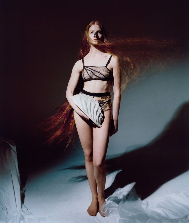 Elegant studio portrait of a red-haired woman in lingerie, holding a ridged clutch bag. Soft lighting highlights her skin tone.