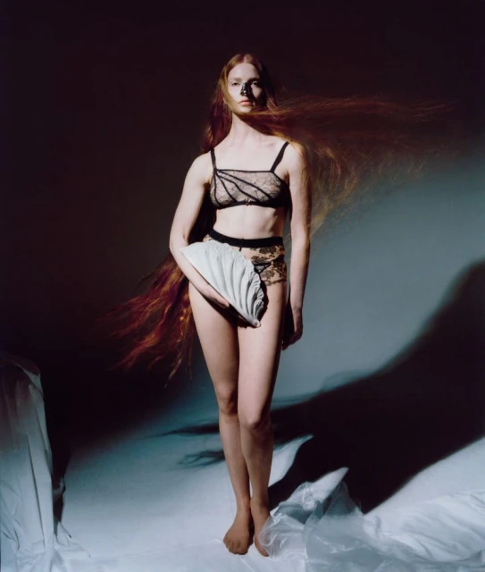 Elegant studio portrait of a red-haired woman in lingerie, holding a ridged clutch bag. Soft lighting highlights her skin tone.