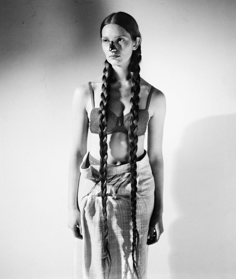 Black and white portrait of a woman with long braids, wearing a bra top and skirt, posing in front of a plain background with shadows.