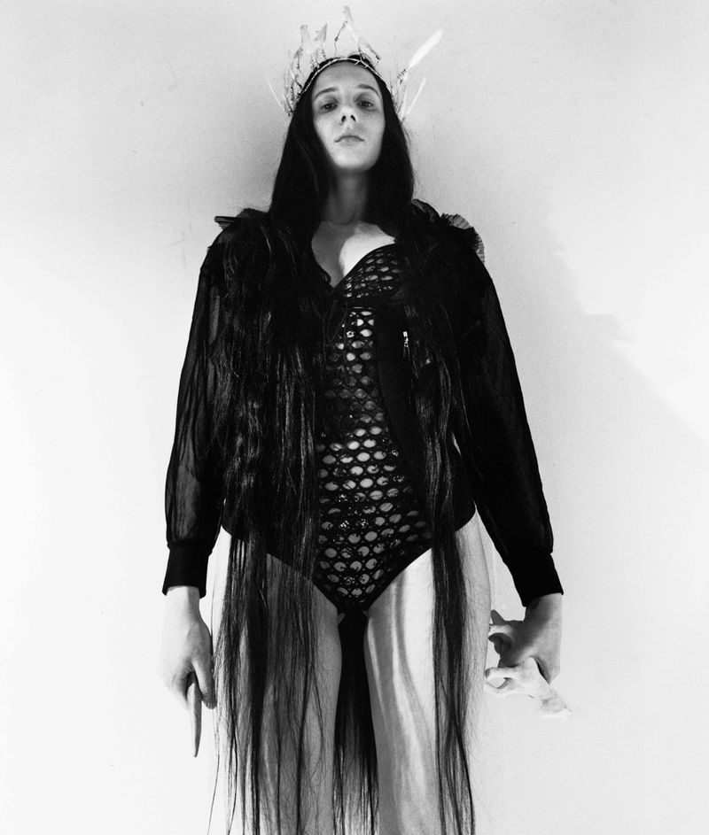 Monochrome portrait of a person wearing a crown and bodysuit with dark wavy hair, posing against a white wall in studio lighting.