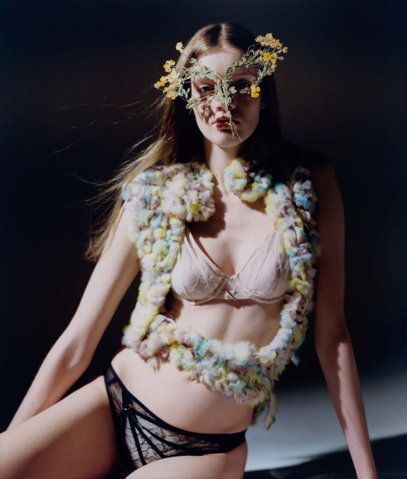 A young woman poses in a bra and underwear, wearing a flower crown and textured vest, against a dark backdrop in a fashion photo.