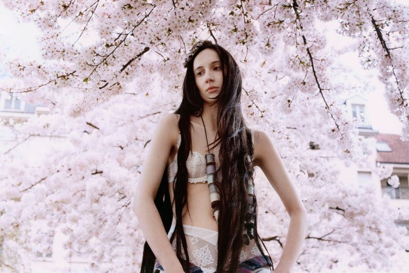 Adult female poses in lingerie with long dark hair, jewelry, and plaid shirt against a cherry blossom tree with a neutral expression.