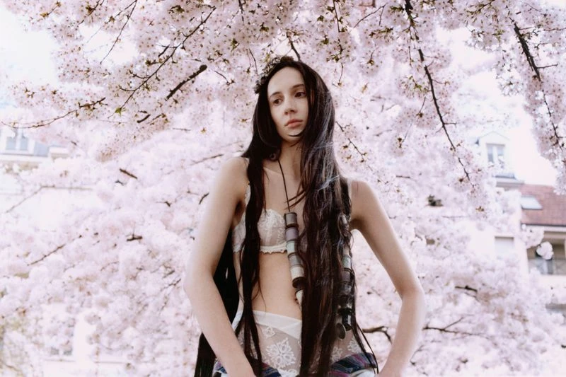 Adult female poses in lingerie with long dark hair, jewelry, and plaid shirt against a cherry blossom tree with a neutral expression.