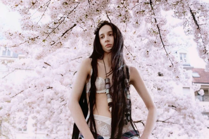 Adult female poses in lingerie with long dark hair, jewelry, and plaid shirt against a cherry blossom tree with a neutral expression.