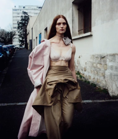 A fashion photo of a tall, thin female model in lingerie and draped coat walking on a city street with buildings in the background.