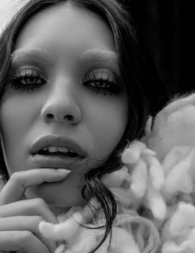 Black and white close-up portrait of Maddie Ziegler with bleached eyebrows, dramatic lashes, and a fluffy textured collar.