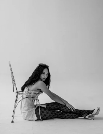 Maddie Ziegler seated on the studio floor leaning against a distressed metal chair wearing patterned tights and high heels.