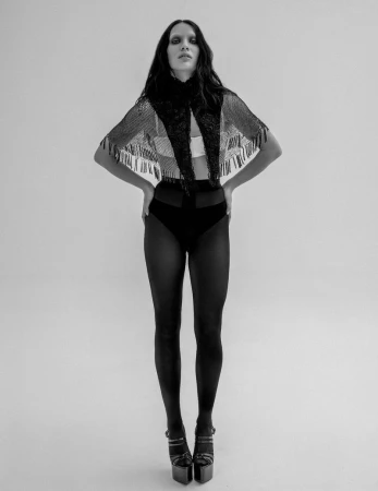 Maddie Ziegler standing hands on hips wearing a black fringed capelet, high-waisted briefs, tights, and platform heels in a studio.