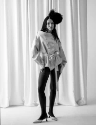 Maddie Ziegler wearing a belted jacket, black tights, and a feathered headpiece against white curtains for DSECTION Magazine.