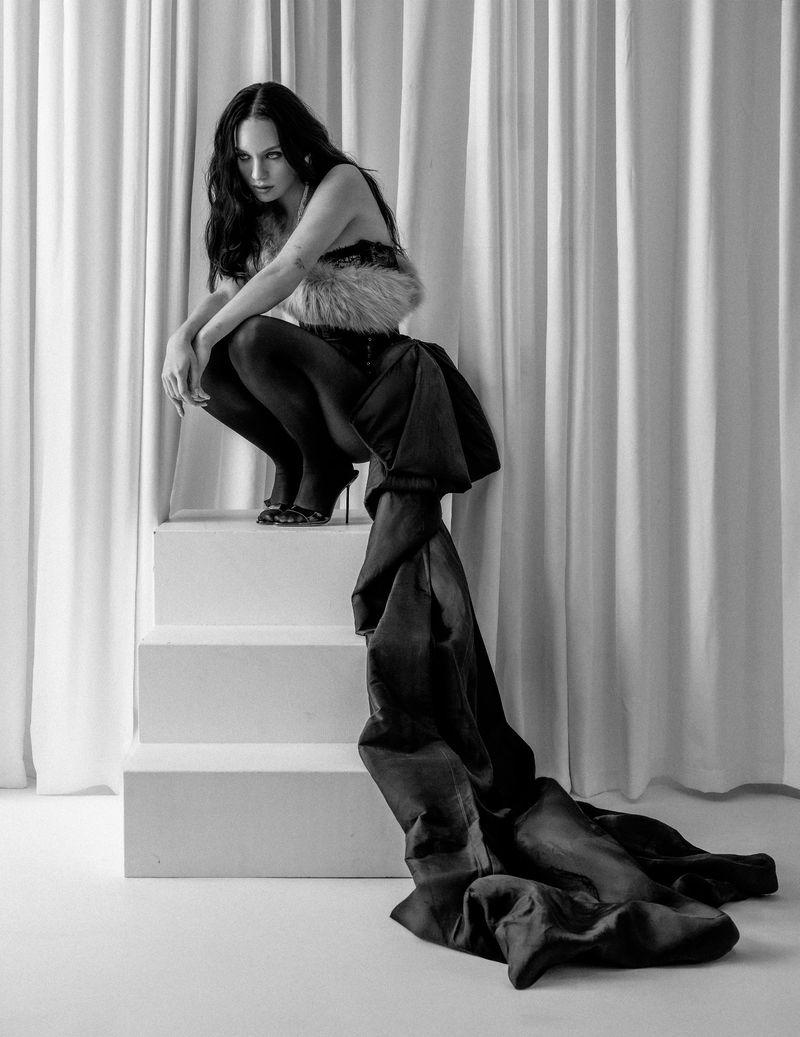 Maddie Ziegler crouching on white steps wearing dark tights and an outfit with a long black fabric train against draped curtains.