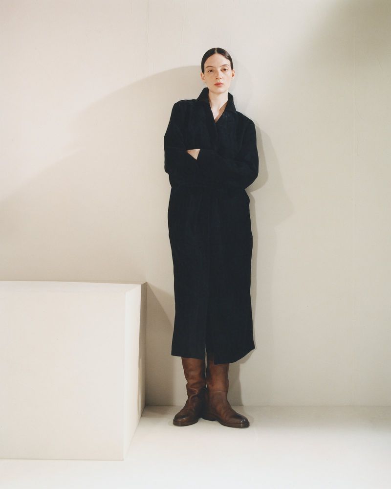 Model Lila Braghero in a long black coat and brown leather boots standing with arms crossed beside a cube prop in a studio setting.
