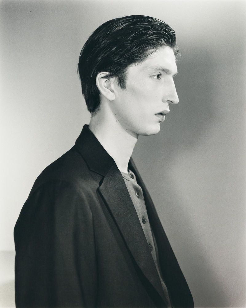 Side profile portrait of a model with slicked-back dark hair wearing a dark tailored blazer against a neutral studio backdrop.