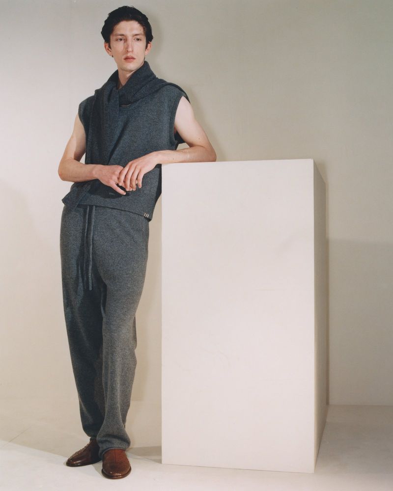 Male model in a sleeveless grey knit top and matching trousers leaning on a white plinth against a pale studio wall.