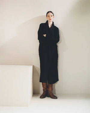 Model Lila Braghero in a long black coat and brown leather boots standing with arms crossed beside a cube prop in a studio setting.