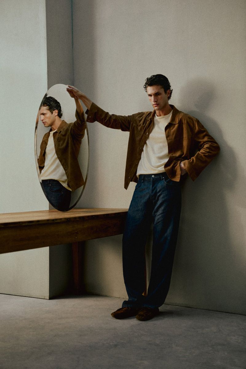 Model Francisco Henriques standing in a brown jacket and wide-leg jeans beside an oval mirror resting on a wooden console table.