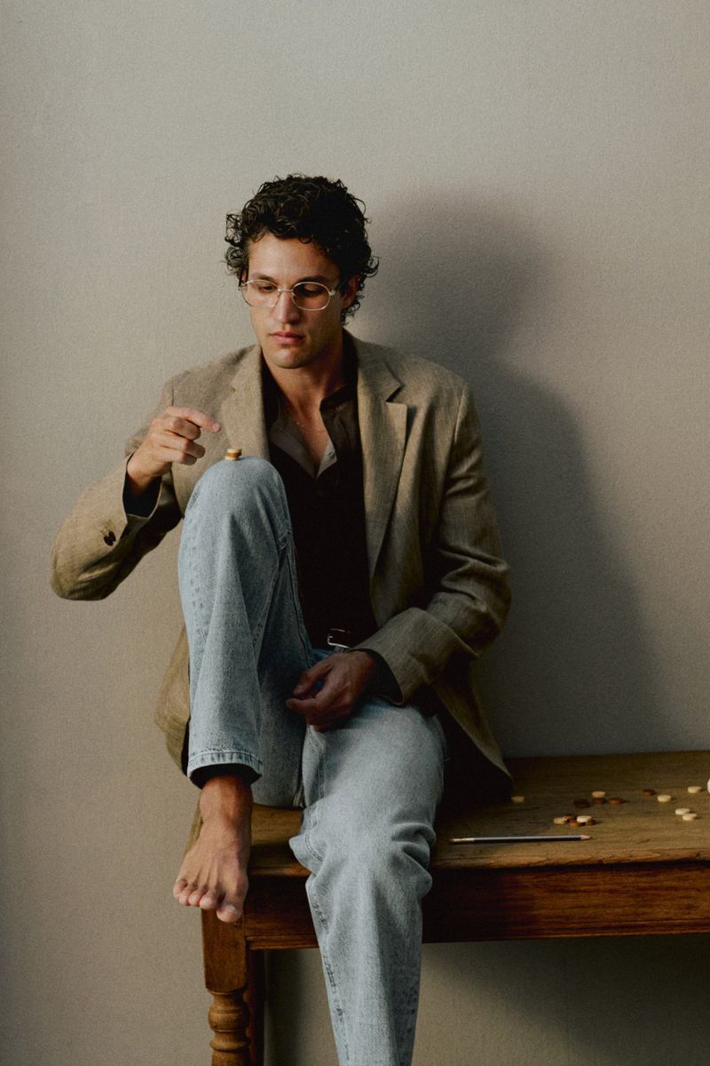 Francisco Henriques wearing a tan blazer and light blue jeans, seated barefoot on a rustic wooden table against a neutral wall.