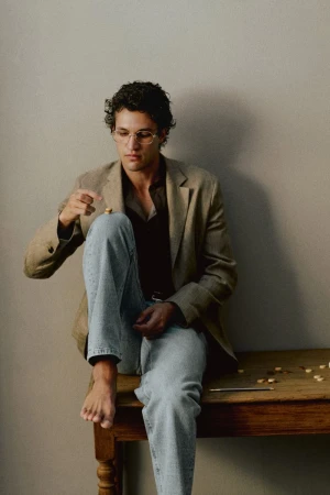 Francisco Henriques wearing a tan blazer and light blue jeans, seated barefoot on a rustic wooden table against a neutral wall.