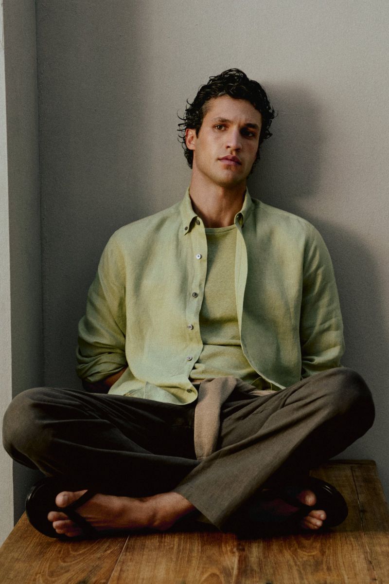 Model Francisco Henriques seated cross-legged in a sage green linen shirt and olive trousers against a neutral wall.