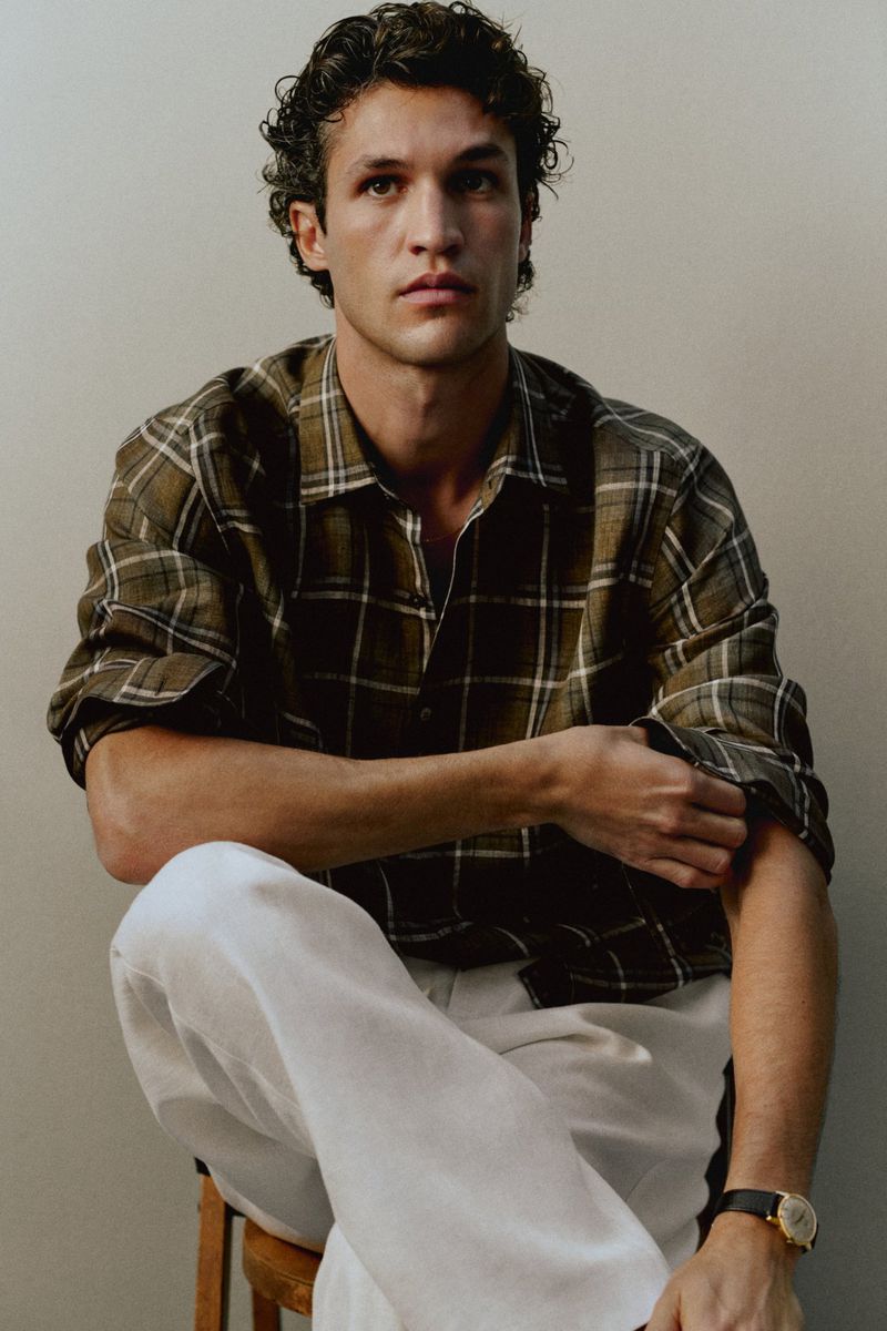 Francisco Henriques wearing a plaid shirt and white trousers seated against a plain neutral wall for Massimo Dutti