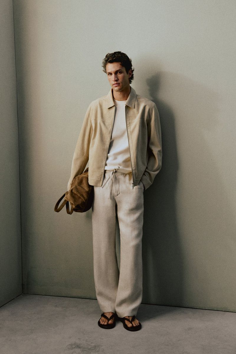 Model Francisco Henriques leaning against a studio wall in a beige jacket, white shirt, cream trousers, and sandals.