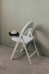 White folding chair holding a stack of notebooks and a small white cup on a saucer beside a plain wall.