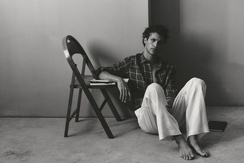 Francisco Henriques sitting barefoot on the floor in a plaid shirt and white trousers beside a wooden chair.
