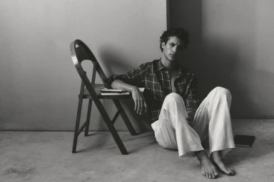 Francisco Henriques sitting barefoot on the floor in a plaid shirt and white trousers beside a wooden chair.