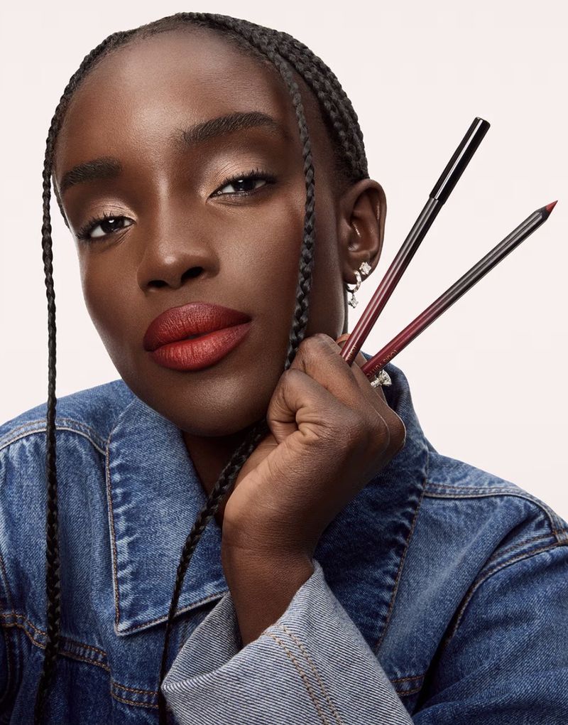 Maty Fall wearing bold red lipstick and a blue denim jacket holding makeup pencils against a pale background.