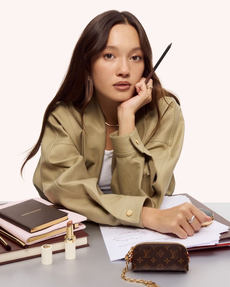 Eileen Gu leaning on a desk with notebooks and a monogram pouch, holding a pencil while wearing a beige leather overshirt.
