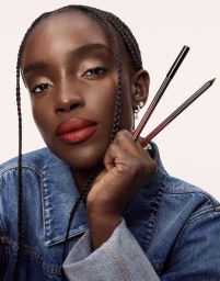 Maty Fall wearing bold red lipstick and a blue denim jacket holding makeup pencils against a pale background.
