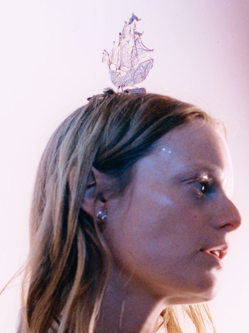 Profile of a woman with light hair and face glitter, wearing a decorative silver ship on her head against a pale pink backdrop.
