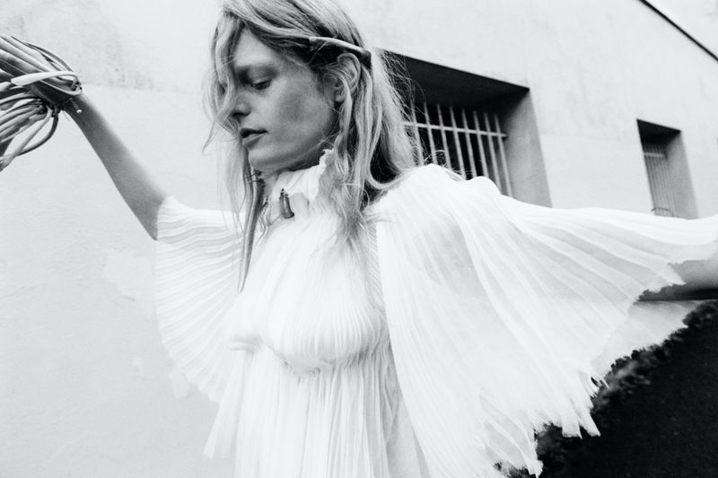Monochrome fashion portrait of a blonde woman in a flowing white dress, holding foliage, against a textured white wall with barred windows.